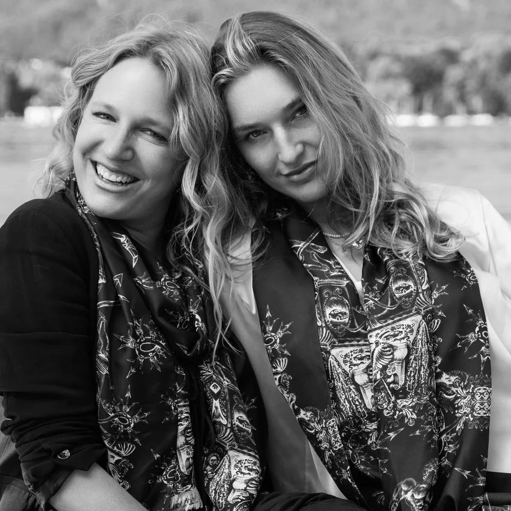 A black and white photograph of two women smiling closely together, both wearing matching Ginny Litscher printed silk scarves
