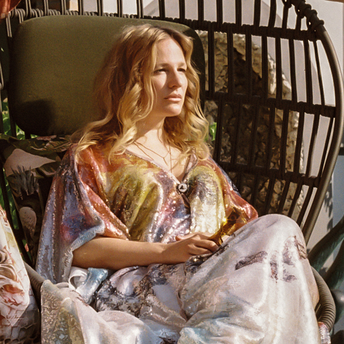 Ginny Litscher in a painted sequin kaftan, seated in a wicker chair